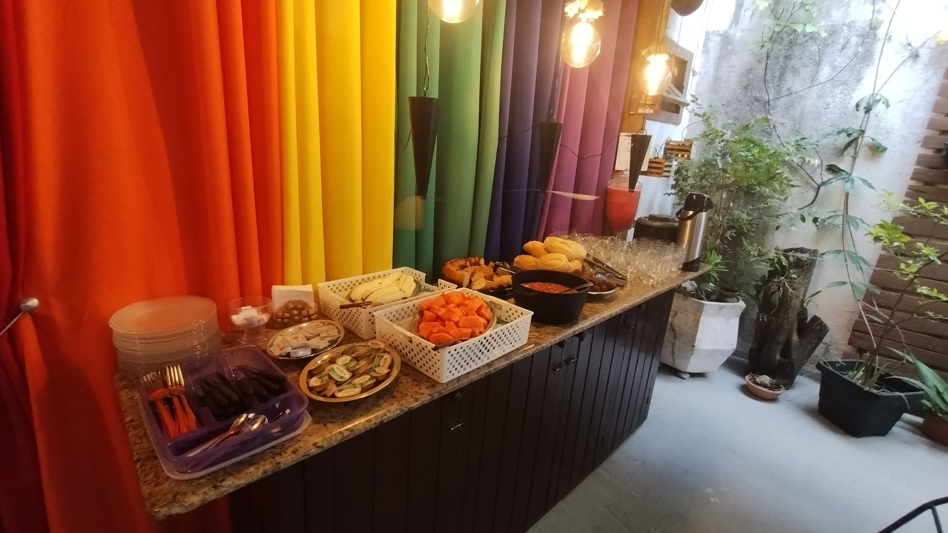 Mesa de buffet com pratos variados ao lado de cortinas laranja e vasos de plantas.