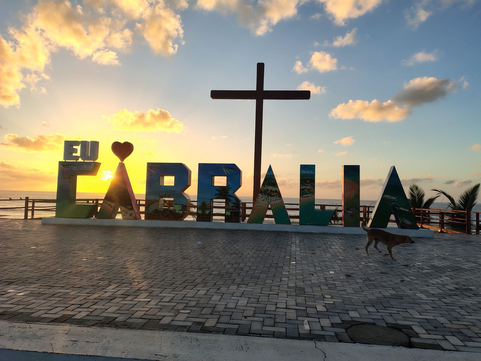 Pôr do sol em Farbala: placa com uma grande cruz e nuvens sobre uma praça pavimentada.