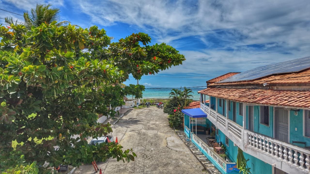 Rua litorânea colorida com árvores, prédios turquesa e vista para o oceano sob um céu azul.
