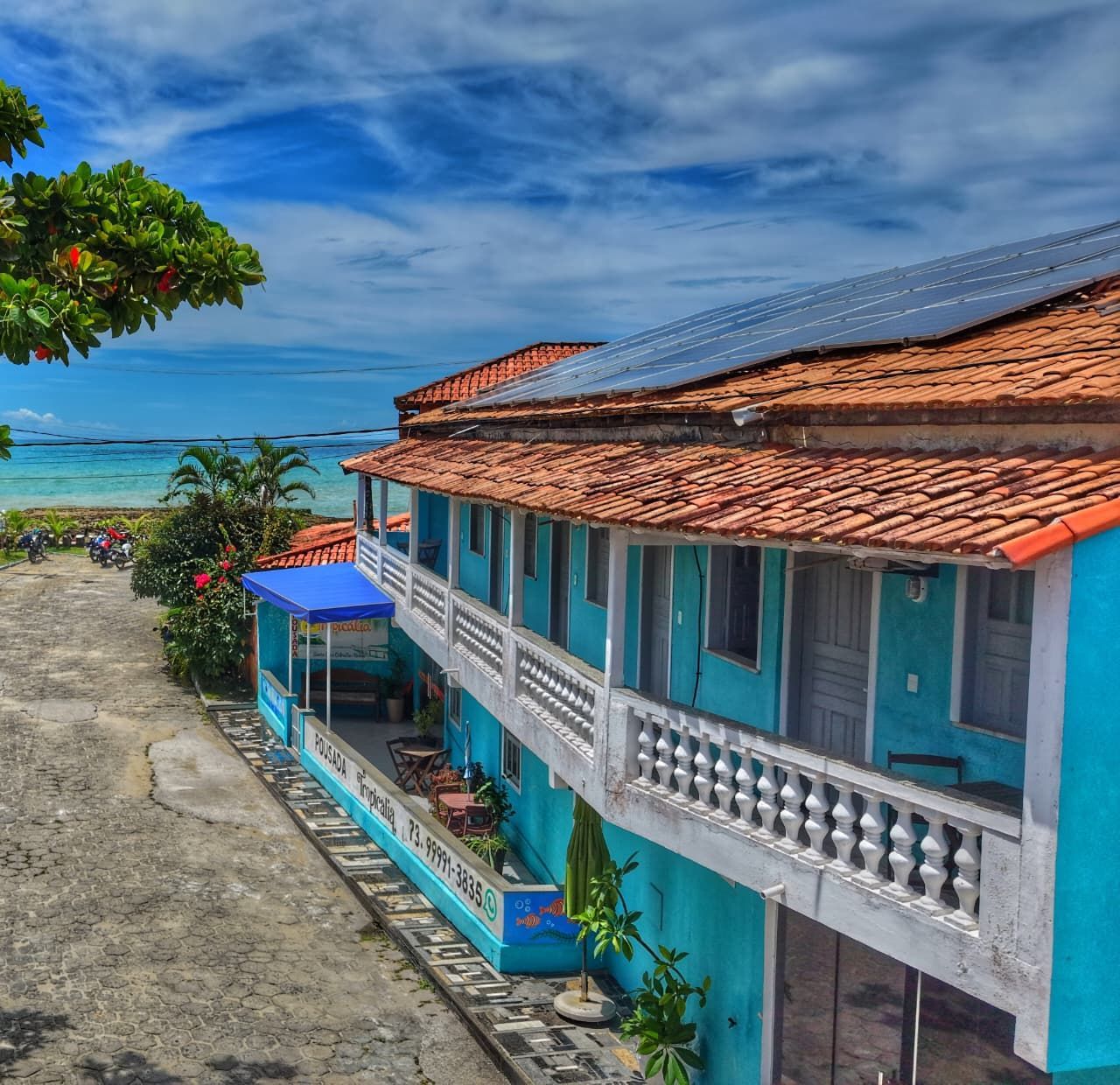 Casa colorida à beira-mar com telhado azul e varanda com vista para uma praia tropical sob um céu claro.