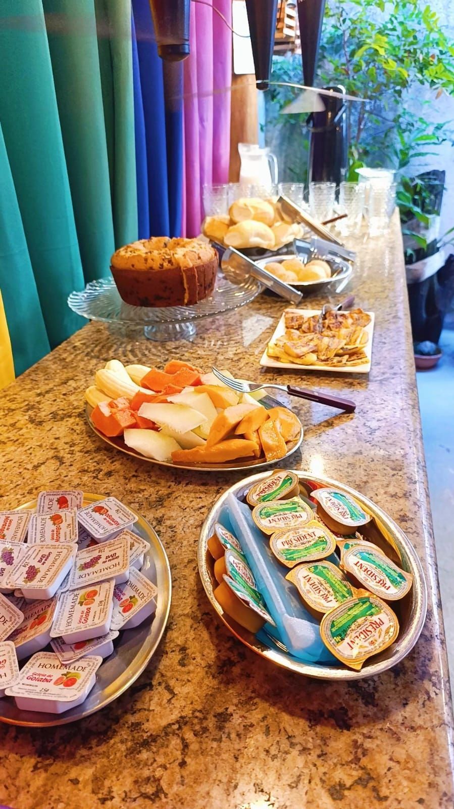 Buffet de café da manhã disposto em um balcão de granito com muffins, frutas, doces e sanduíches variados, protegido por cortinas coloridas.
