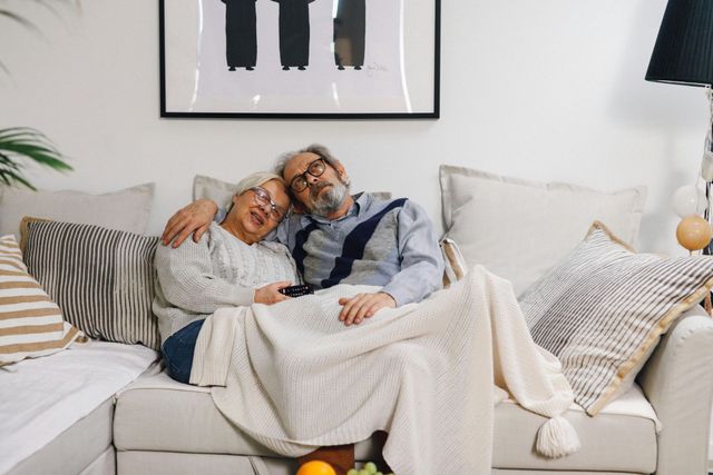 An elderly couple is laying on a couch under a blanket.