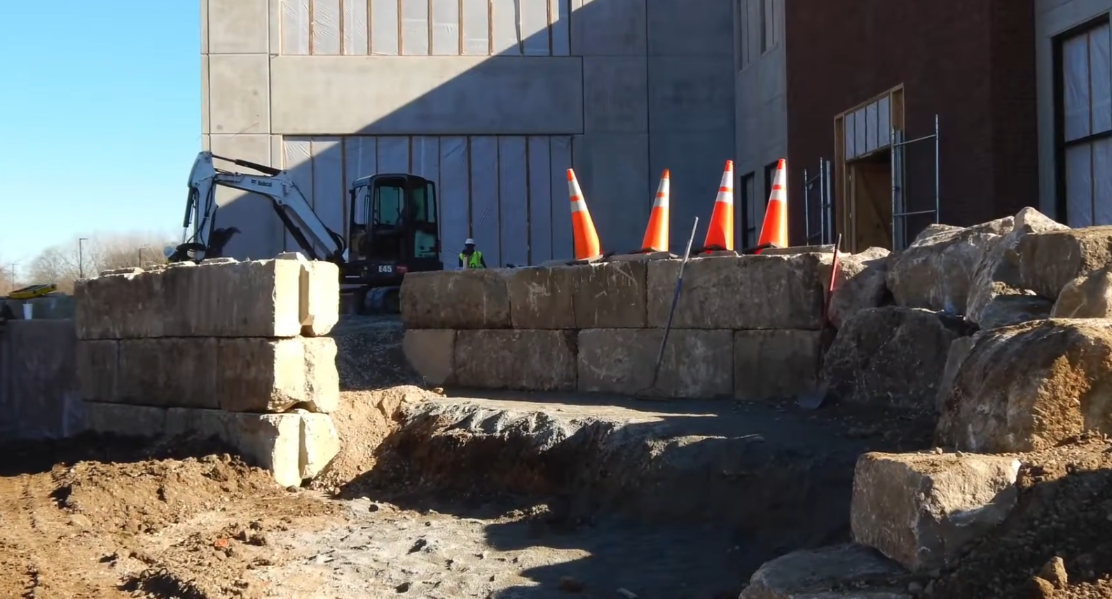 Construction site with stacked hay bales, orange cones, and a blue skid steer by a building wall