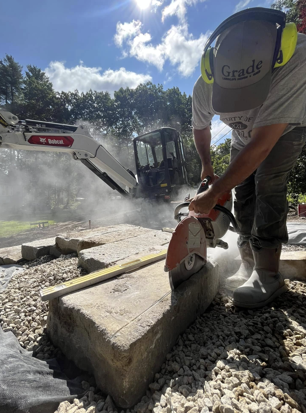 Worker using a cutoff saw to cut a concrete block on a gravel site, with a skid steer spraying water nearby.