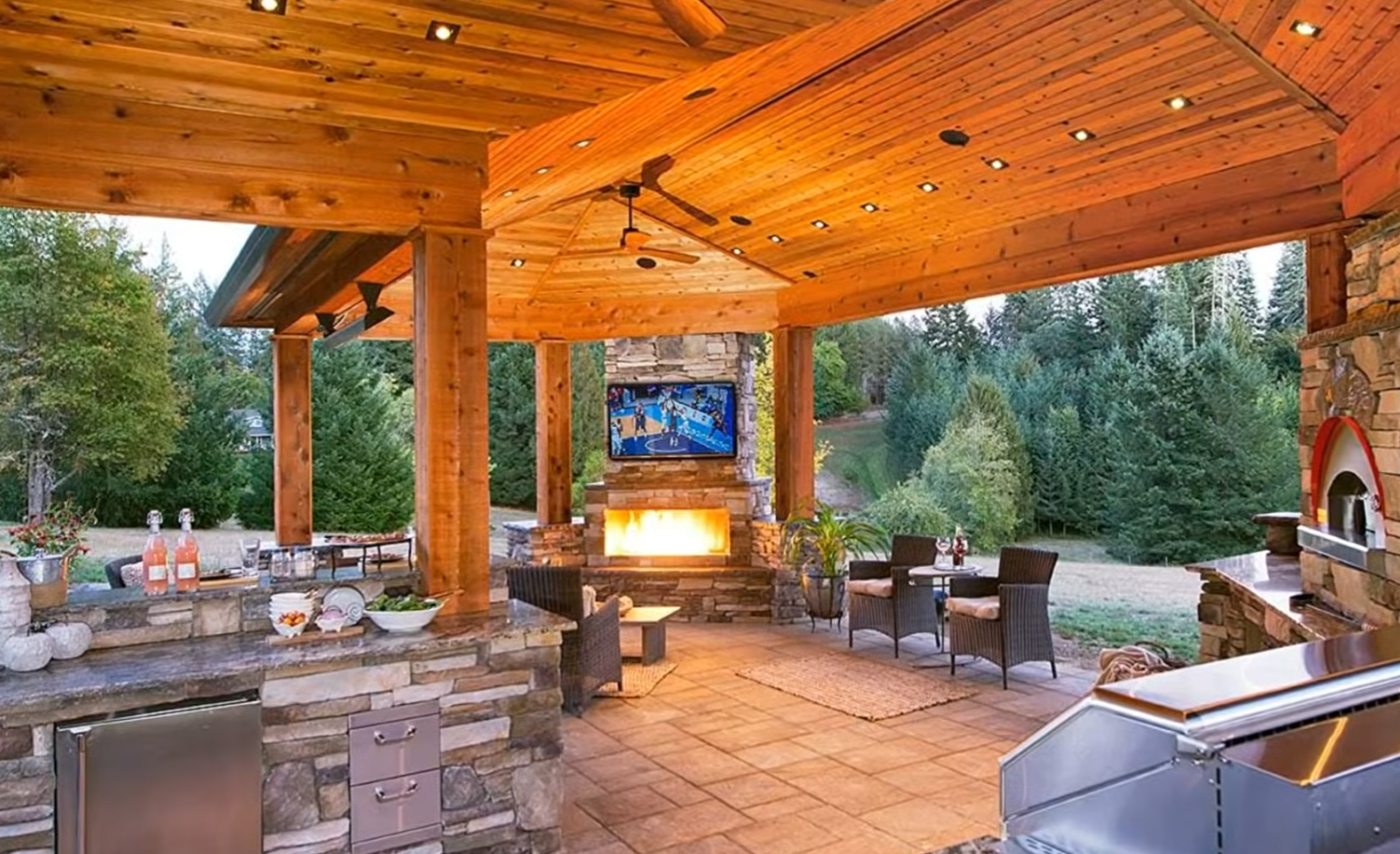 Covered outdoor patio with stone fireplace, wood ceiling, and seating overlooking trees at dusk