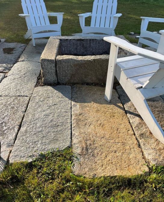 Stone fire pit surrounded by white Adirondack chairs on a patio lawn.