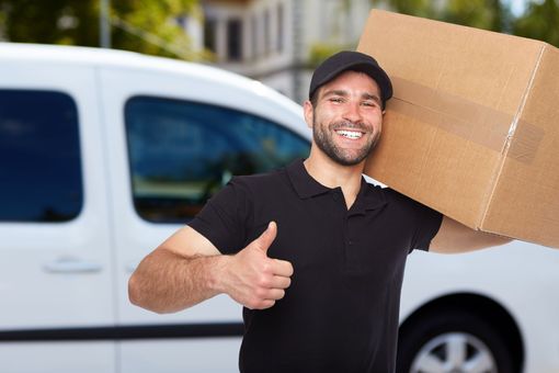 A delivery man is holding a cardboard box on his shoulder and giving a thumbs up.