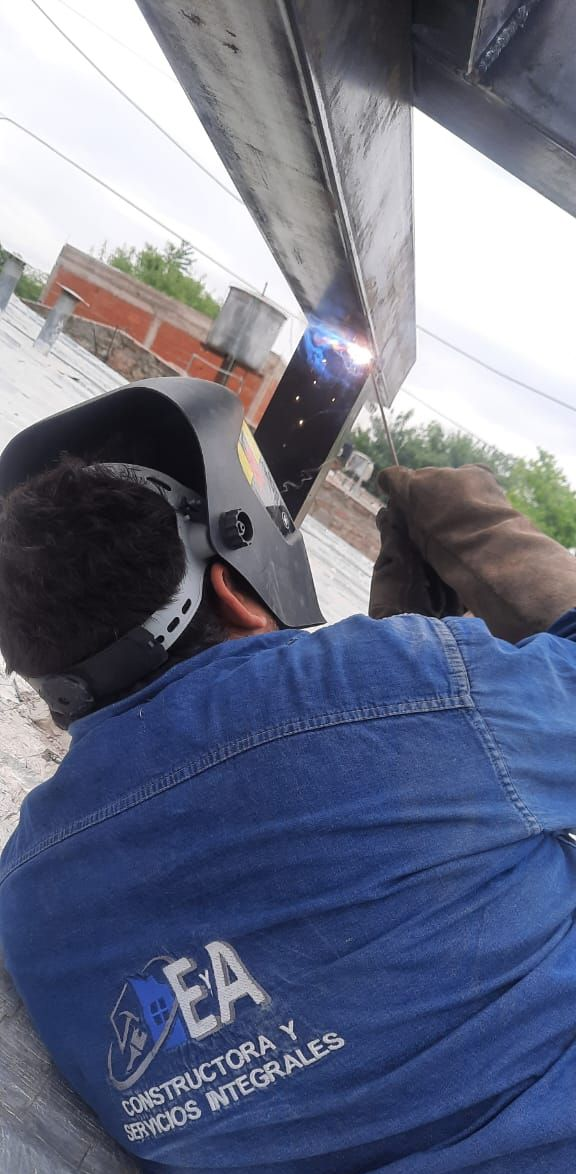 Un soldador con máscara y guantes, suelda una viga de metal al aire libre.