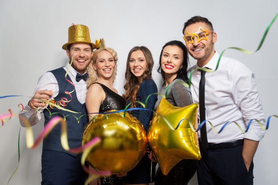 Five people in party hats and festive glasses posing with gold balloons and confetti