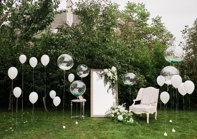 Outdoor wedding setup with white balloons, clear chairs, and a white arch in a garden setting