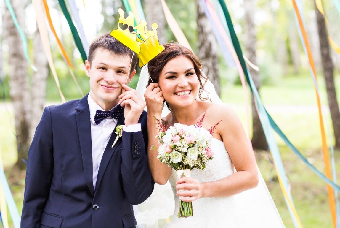 Smiling bride and groom outdoors, holding a bouquet and a gold crown at a wedding ceremony