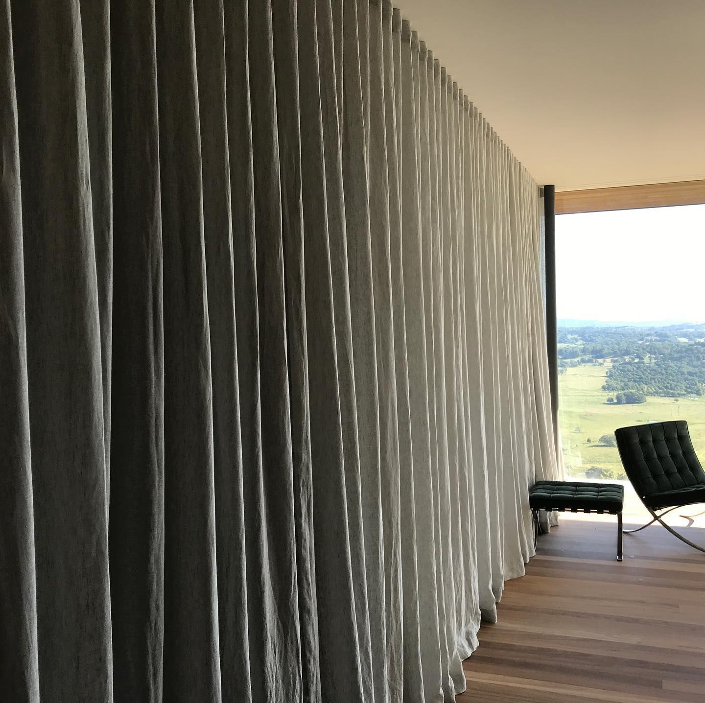 Luxurious floor-to-ceiling grey ripple fold curtains installed in a Byron Bay contemporary home, featuring a textured heavyweight fabric with slight puddle hem, timber flooring, and panoramic rural views through full-height windows
