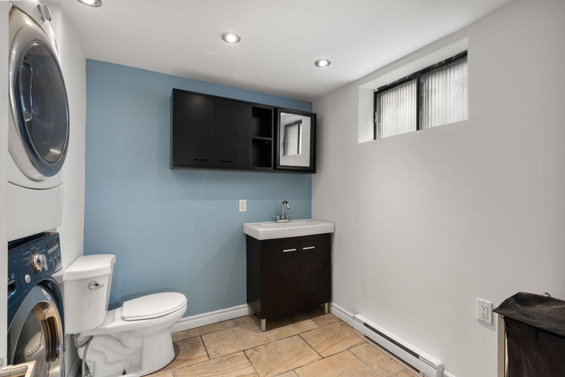 Une petite salle de bains avec toilettes, lavabo, lave-linge et fenêtre. Les murs sont bleus et blancs.