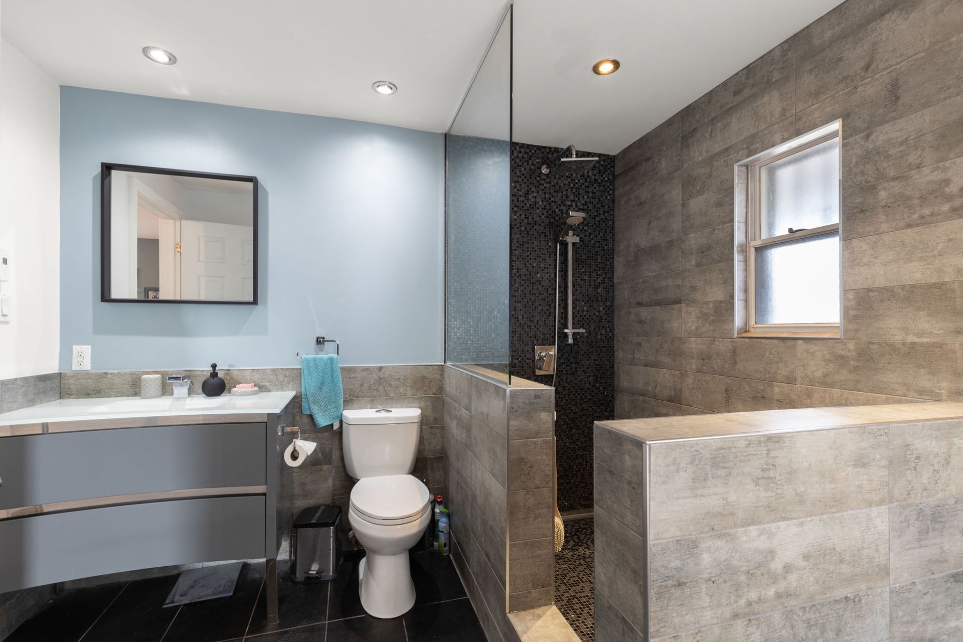 Salle de bains moderne aux tons gris et bleus, douche, toilettes, lavabo et fenêtre.