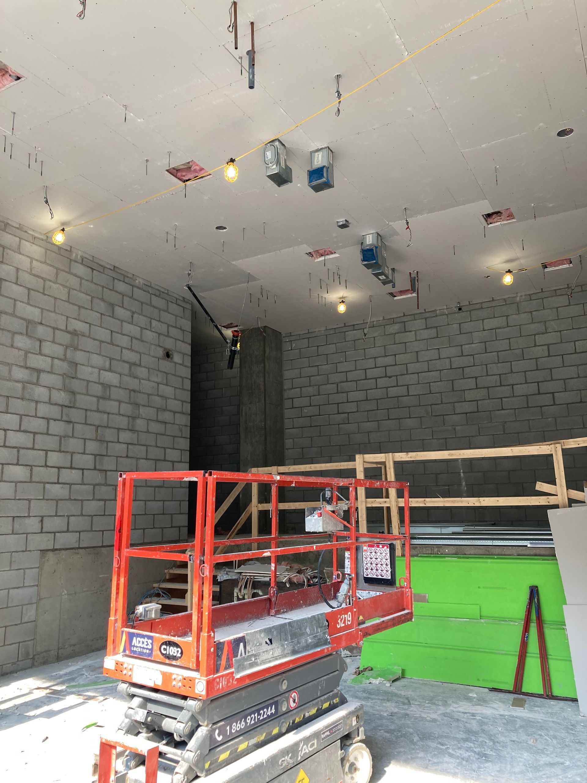 Chantier : nacelle élévatrice rouge, murs en parpaings, éclairage au plafond, câblage apparent.