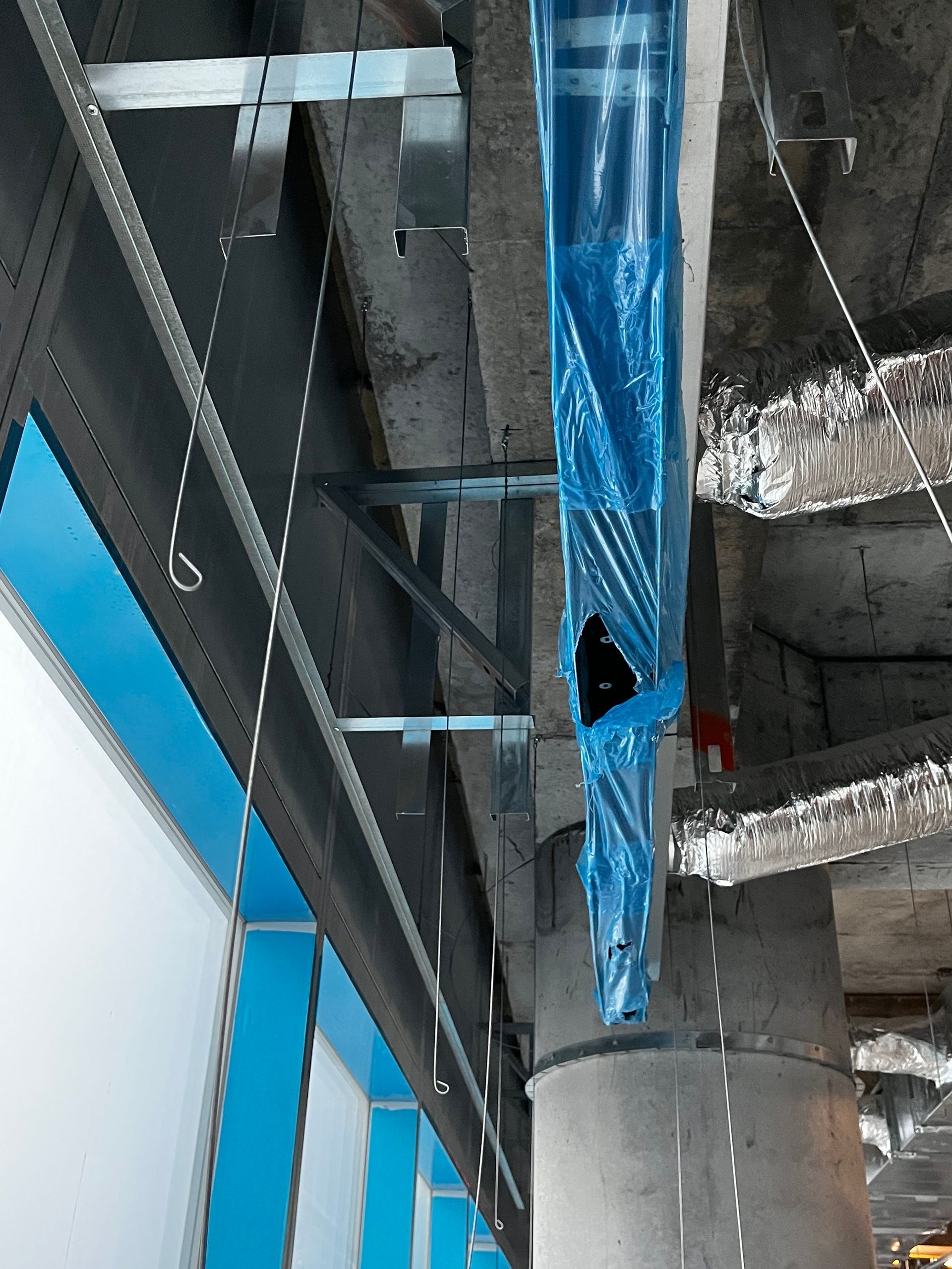 Intérieur d'un chantier de construction avec des poutres apparentes, des tuyaux enveloppés de plastique bleu et une fenêtre à vitre bleue.