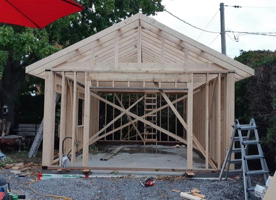 Abri en bois en construction ; charpente terminée, chevrons de toit visibles ; outils et échelle présents.
