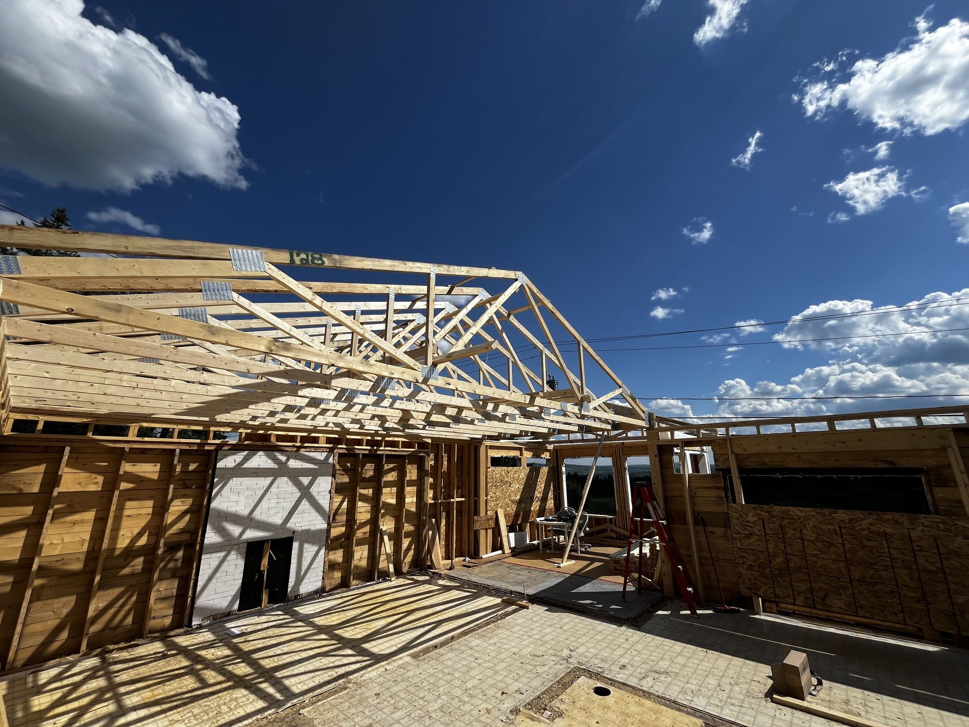 Chantier : charpente en bois d'une maison sous un ciel bleu nuageux. On aperçoit la charpente du toit et les montants des murs.