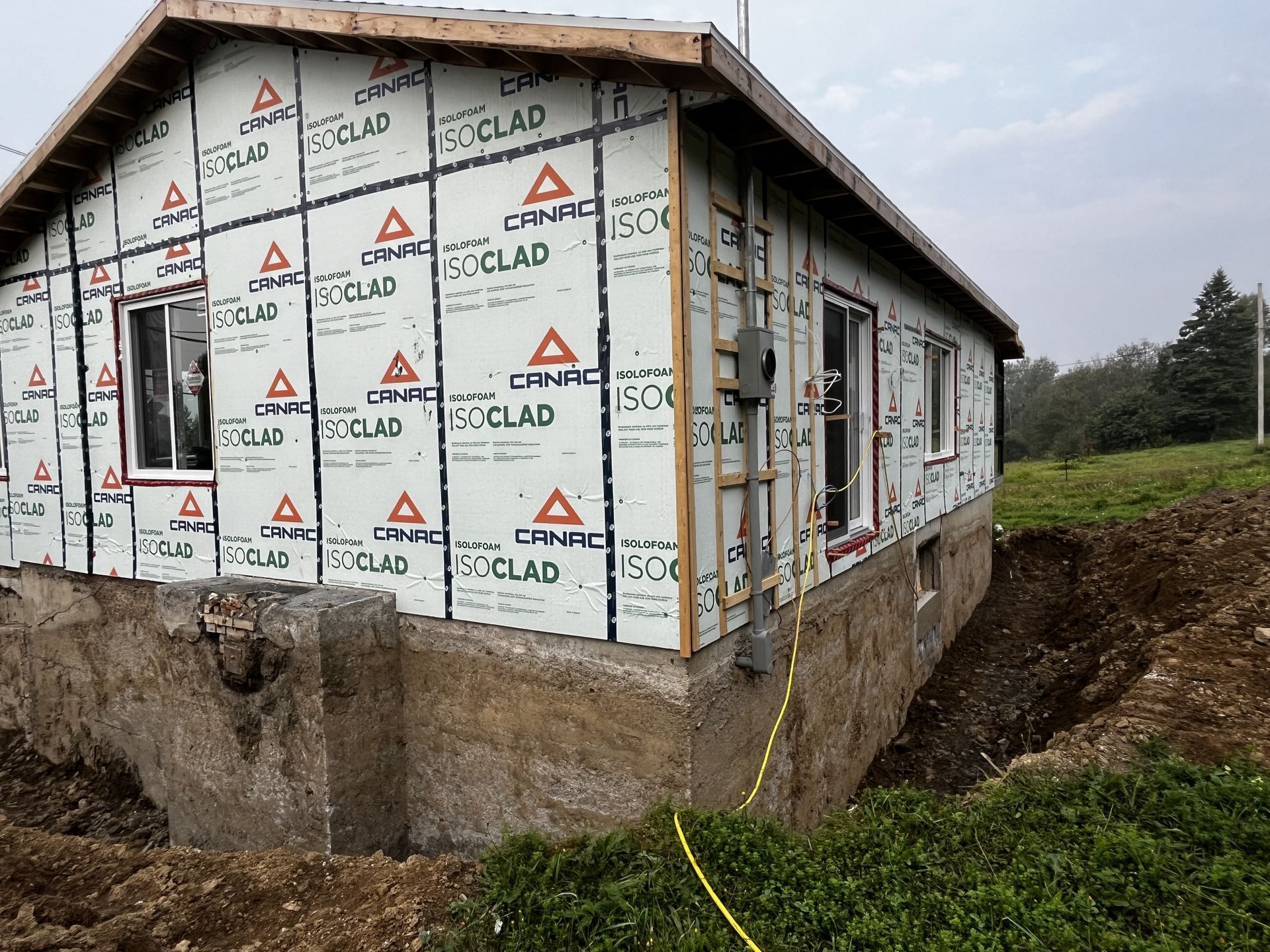 Maison en construction, vue extérieure. Barrière bleue résistante aux intempéries, fenêtres, fondations en béton brut, terre.