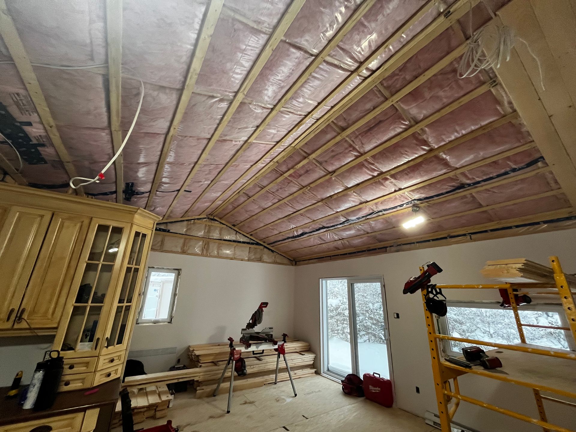 Intérieur d'une pièce en construction avec charpente apparente, isolation rose et vue sur un paysage enneigé à travers une porte vitrée.