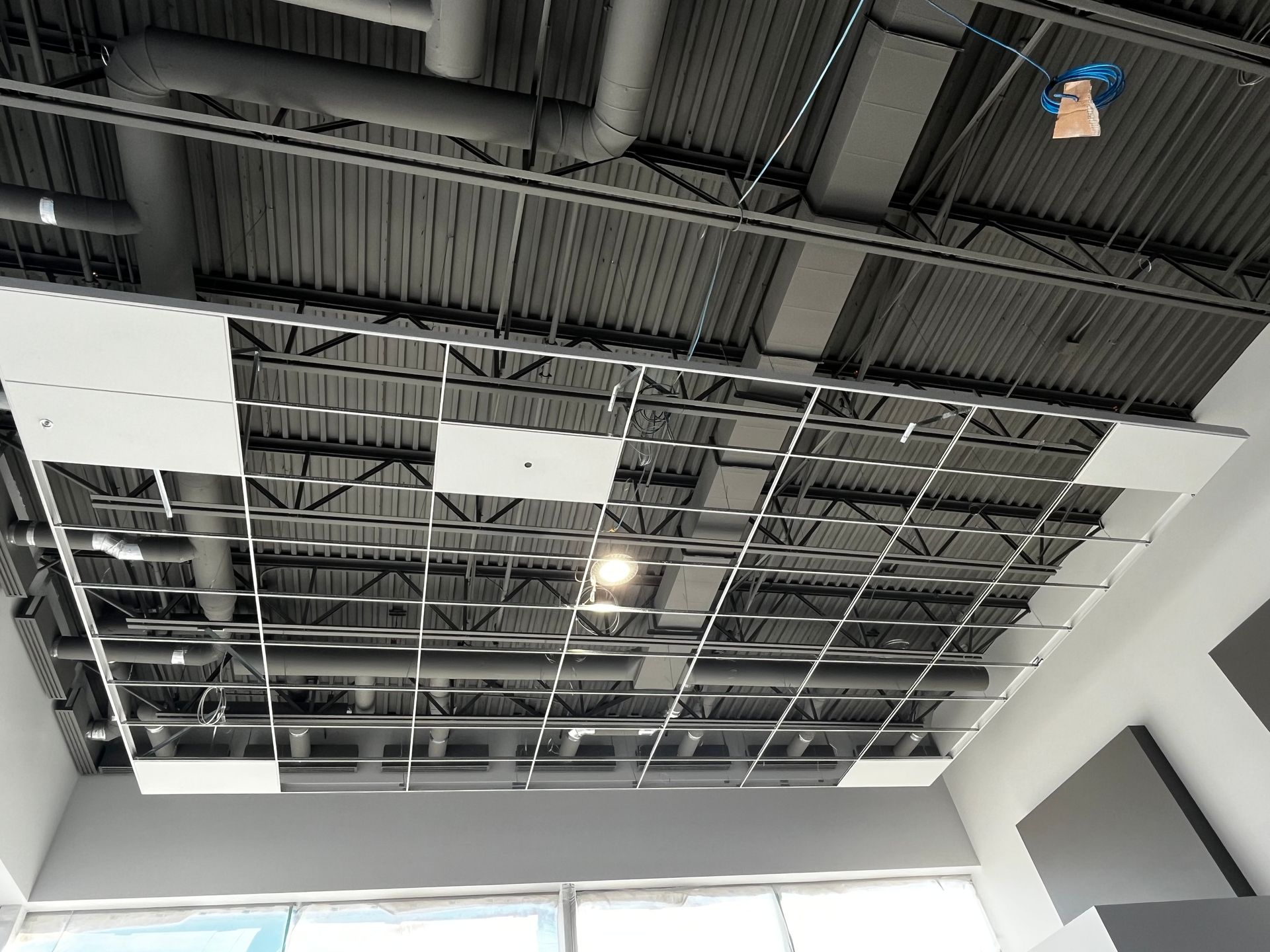 Vue du plafond : Grille métallique avec dalles acoustiques, tuyaux et éclairage dans un espace commercial inachevé.