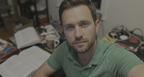 Man in a green polo taking a selfie at a cluttered desk with open books, papers, and coffee mugs