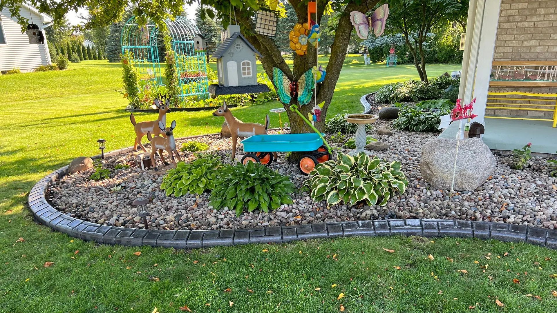 Front yard garden bed with stone border, colorful flowers, birdhouse, and small bench beneath a tree