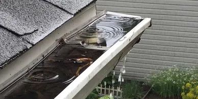 Overflowing rain gutter filled with water and debris, with rain cascading over the edge.