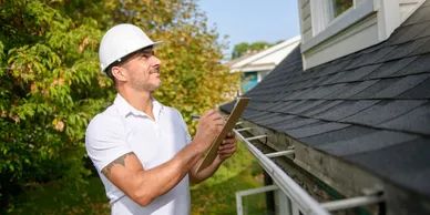 Man in hard hat inspecting roof with a clipboard outdoors.