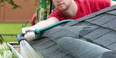 A man in a red shirt uses a hose to clean a gutter on a roof.