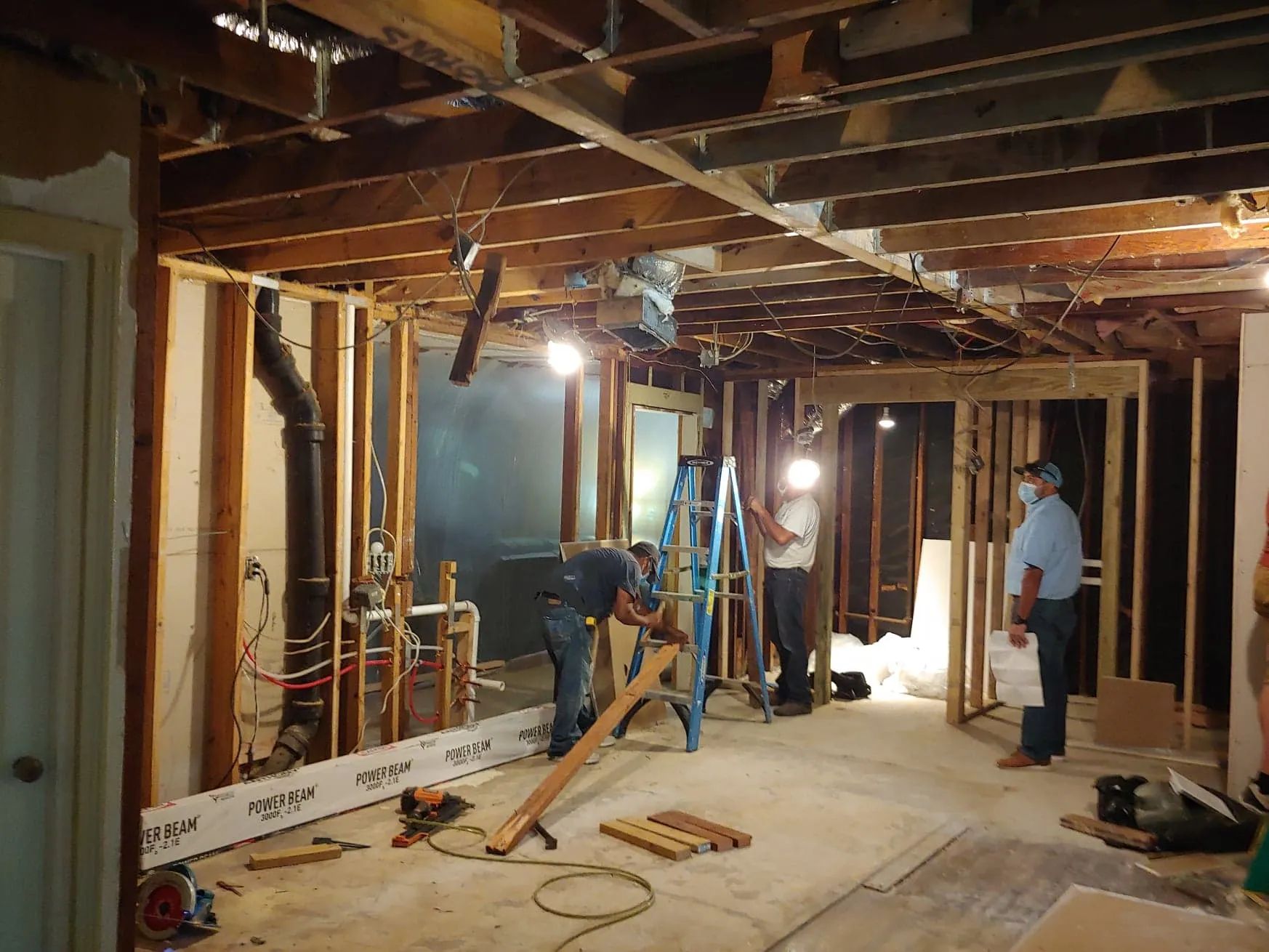 Basement under construction with exposed framing, workers, tools, and debris on the floor.