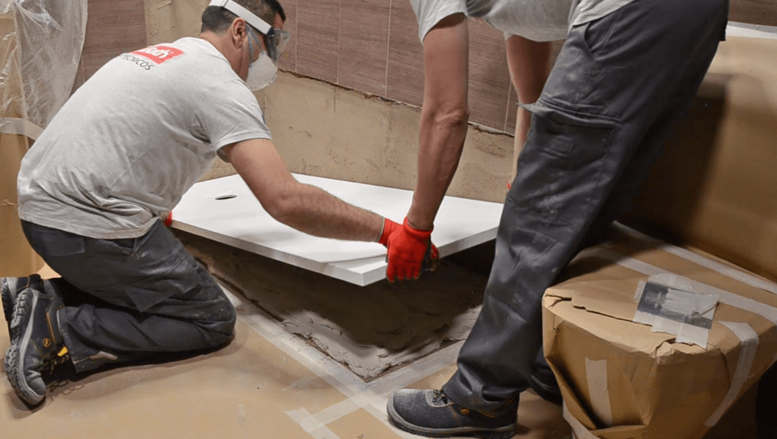 Dos trabajadores instalando una baldosa blanca en una obra; uno arrodillado, el otro de pie, sosteniendo la baldosa.