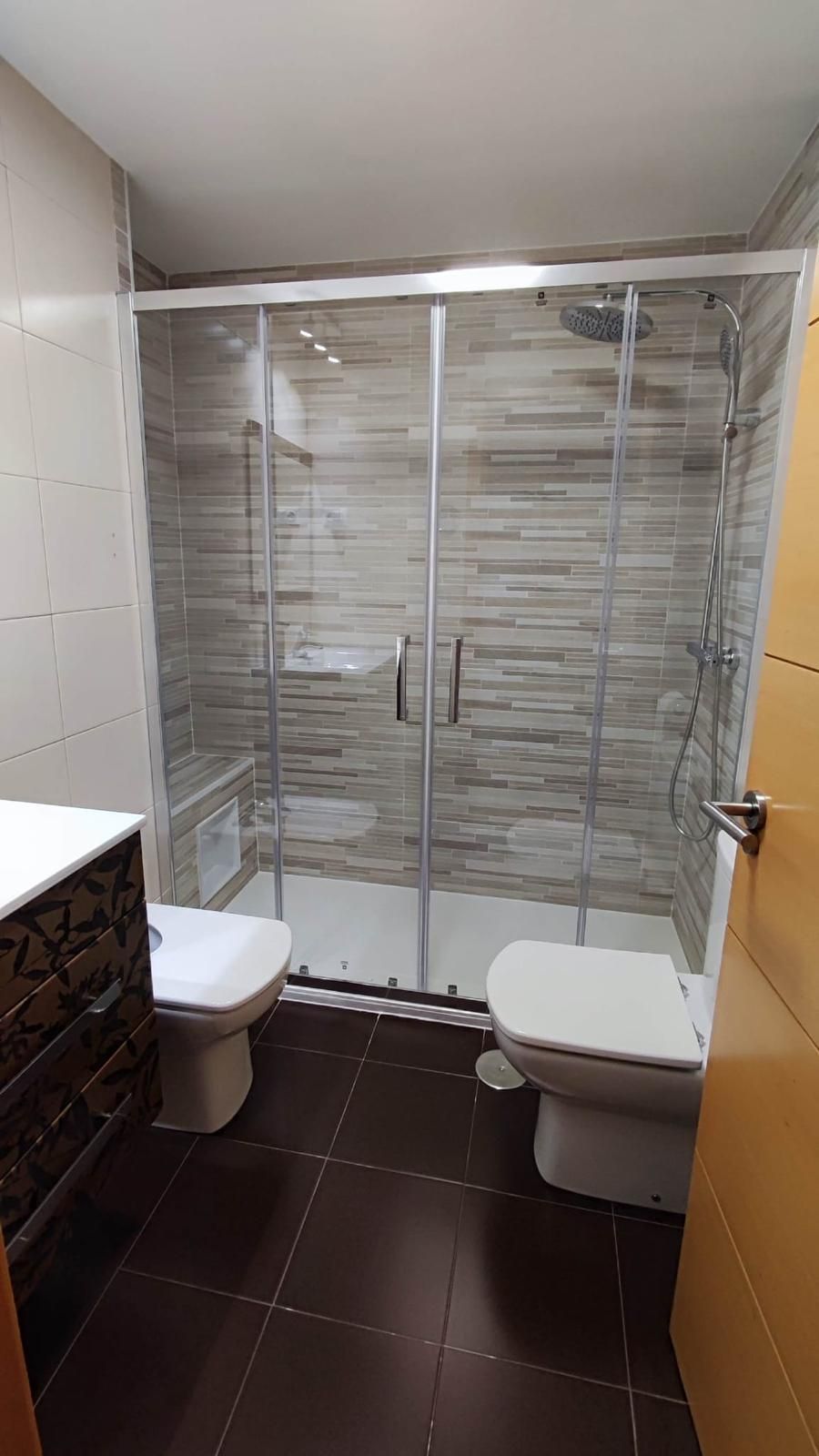 Baño con ducha de cristal, dos inodoros y baldosas de color marrón oscuro en el suelo.