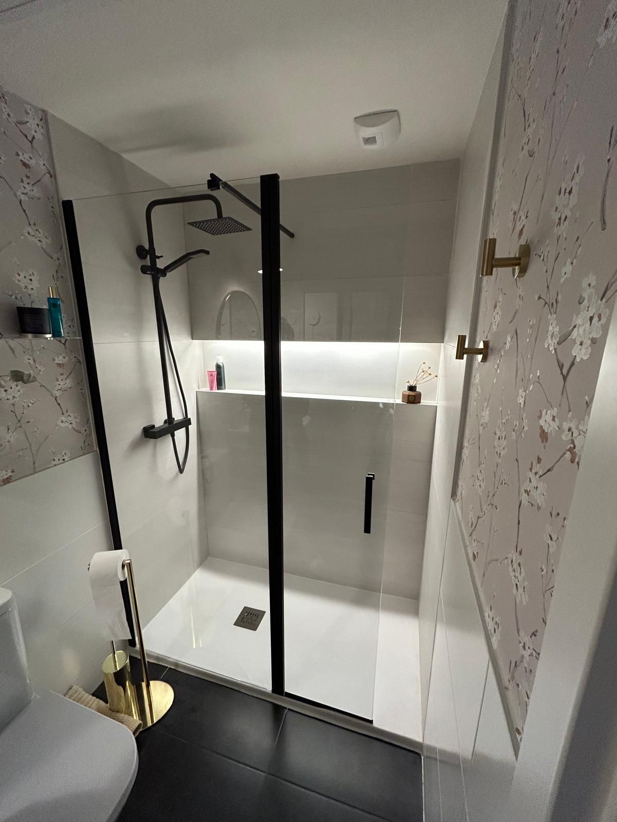 Cabina de ducha moderna con grifería negra y paredes blancas, estante integrado con iluminación y papel pintado estampado.