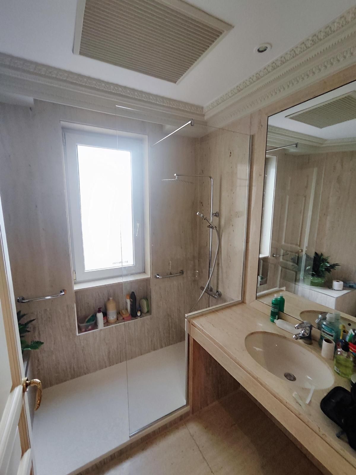 Baño con ventana, ducha y lavabo. Paredes de azulejos beige, espejo, techo blanco y varios artículos de aseo en la encimera y en el hueco de la ventana.
