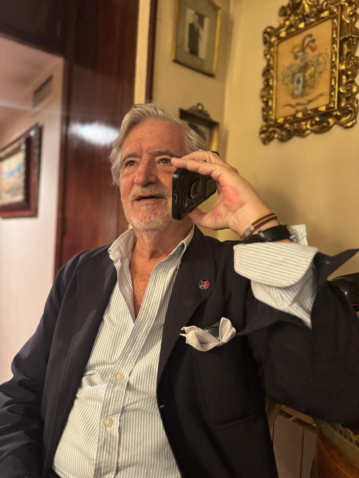Hombre mayor con chaqueta azul hablando por teléfono, en el interior, cerca de una obra de arte con marco dorado.
