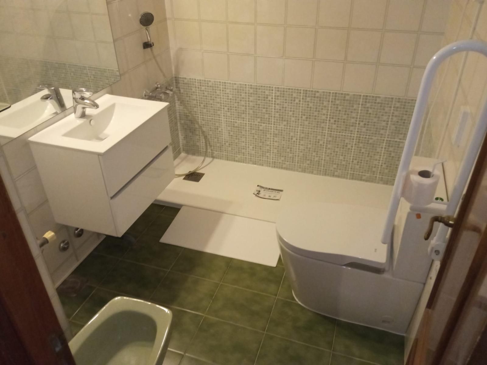 Baño con inodoro, lavabo y barra de seguridad adaptados. Azulejos verdes y blancos, suelo de ducha color crema.