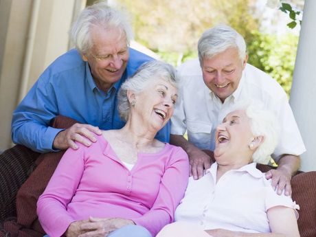 Quattro anziani sorridenti si rilassano insieme in casa, ridendo e stando vicini su un divano.
