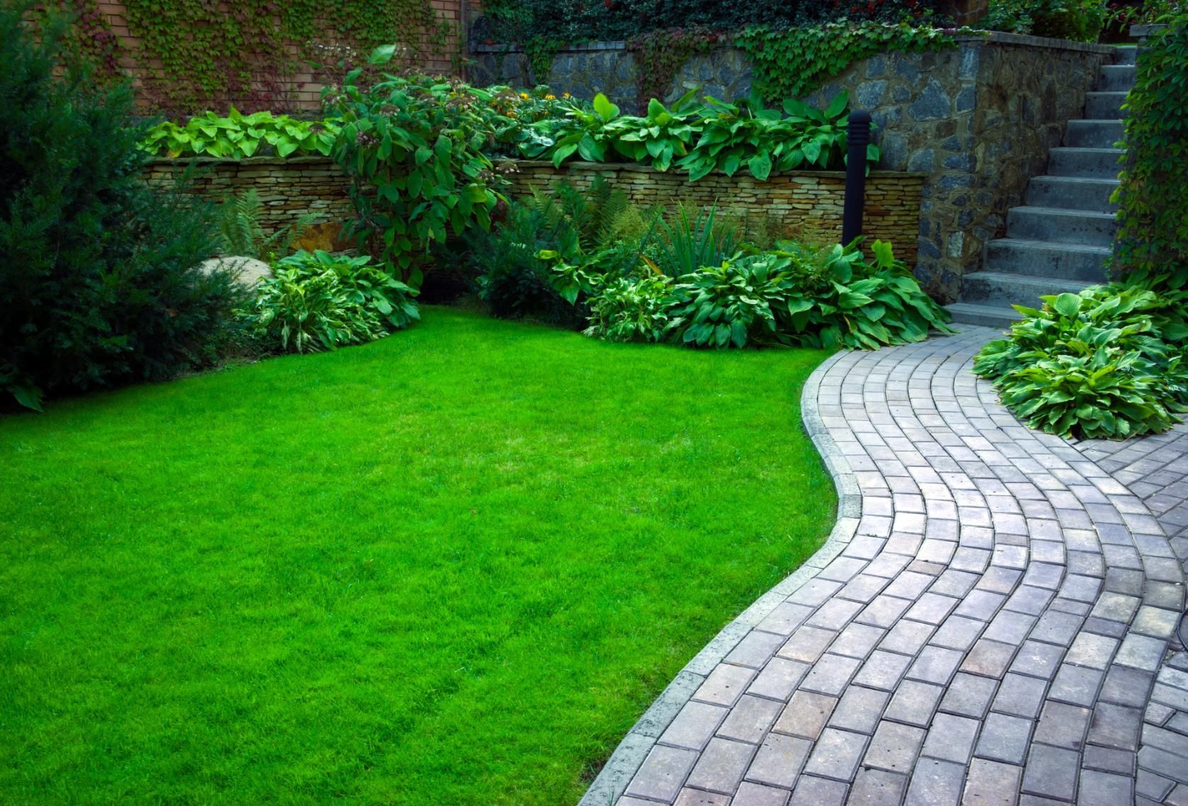 Green lawn and curving stone path in a garden with plants, brick wall, and stairs.