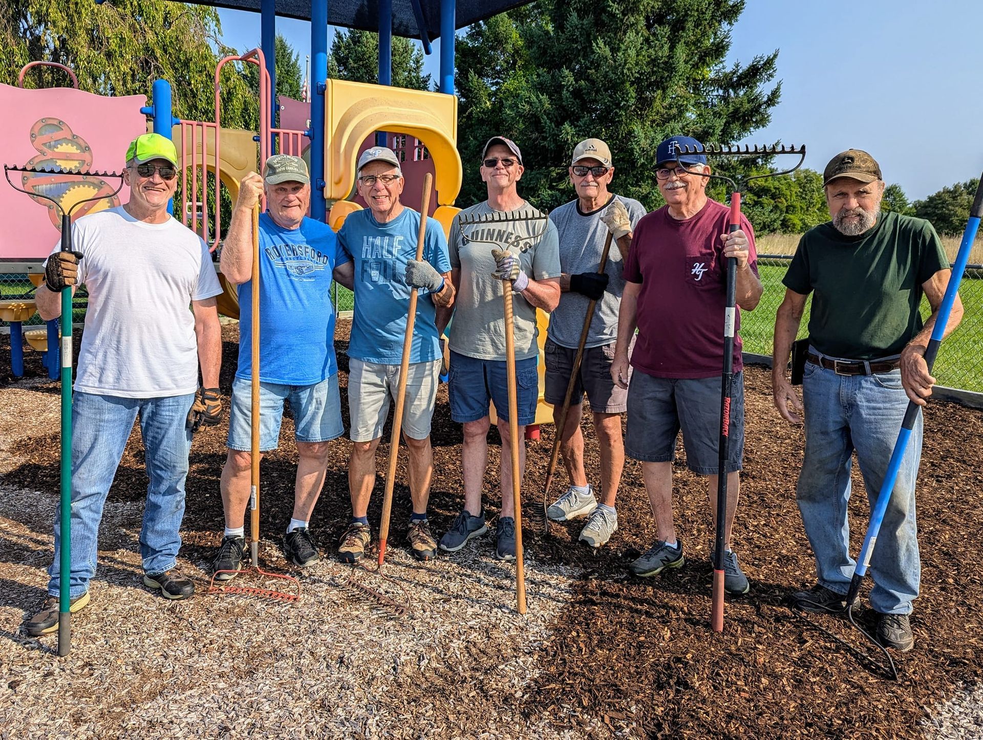 A group of men standing in front of a playground holding gardening tools.
