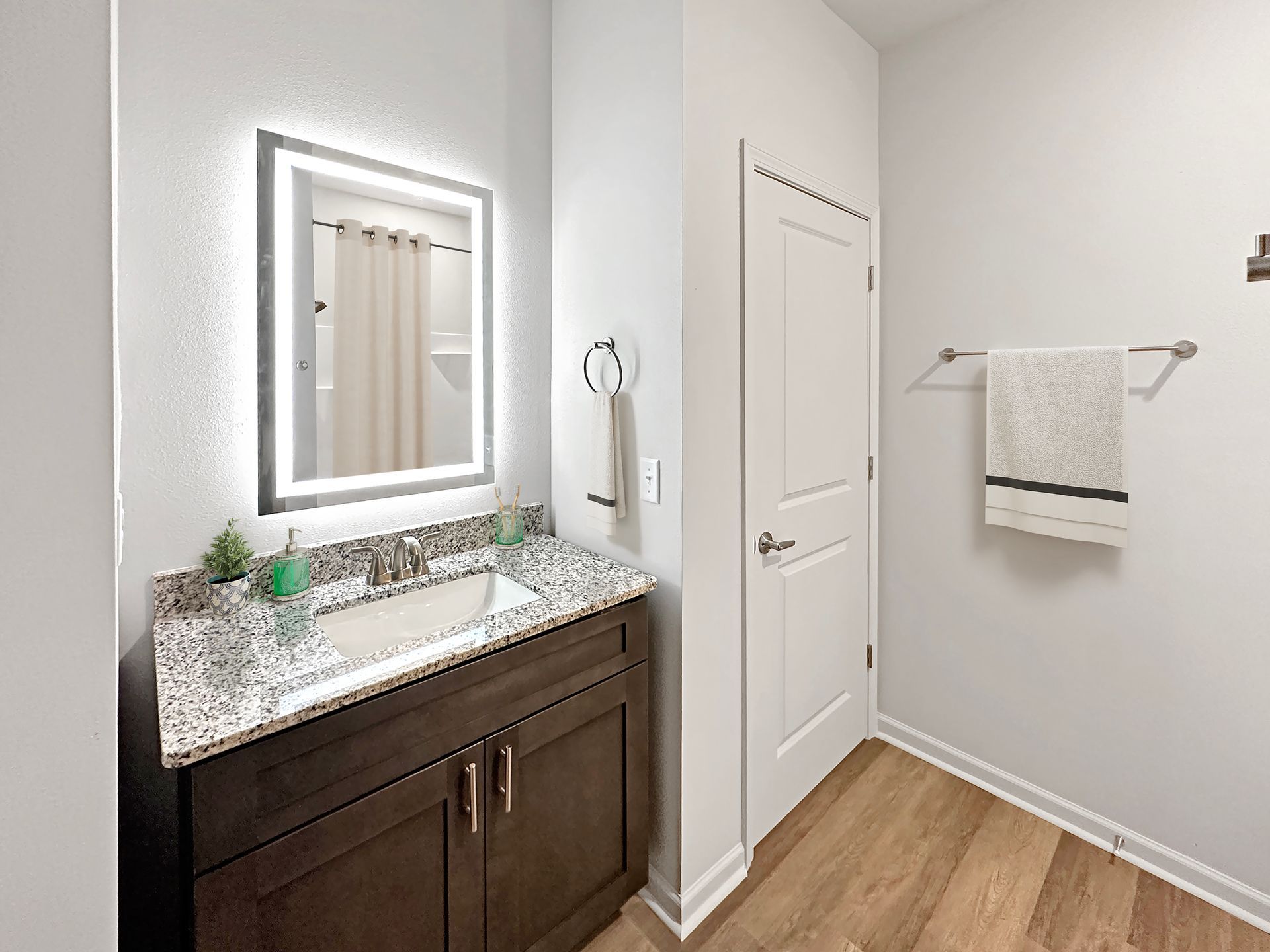 Small bathroom vanity with lit mirror, granite counter, white door, towel rack, and wood-look floor.