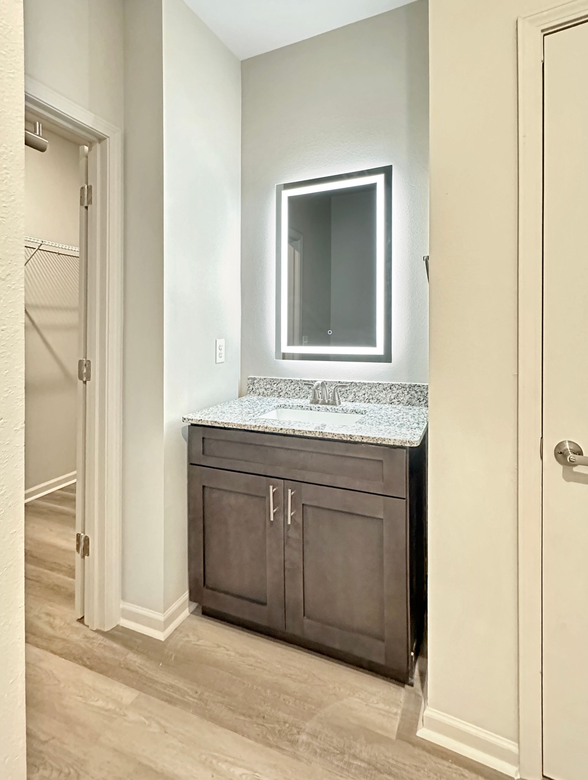 Modern bathroom vanity with dark wood cabinet, speckled countertop, and illuminated mirror in a light hallway-style space
