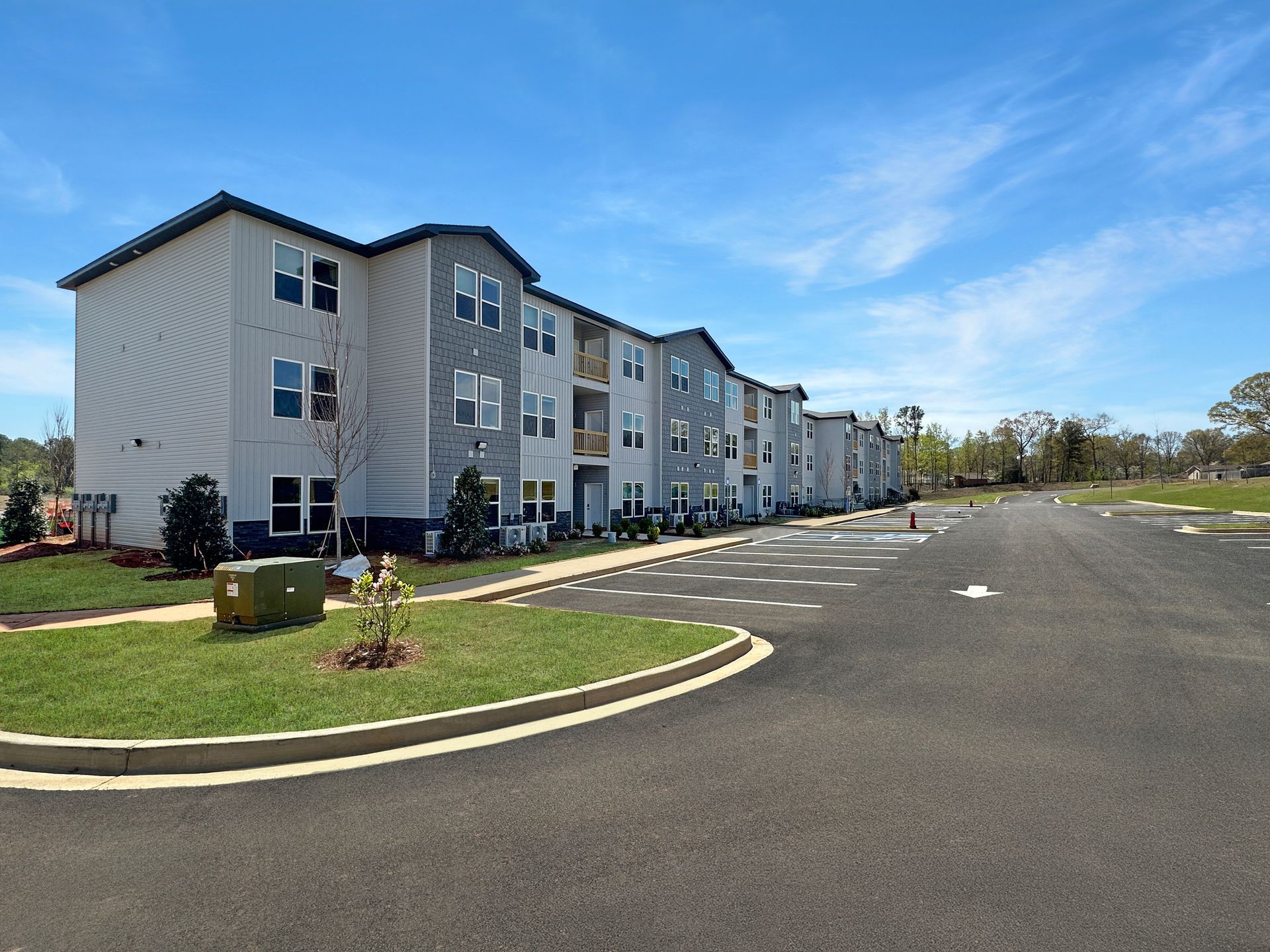 Apartment complex with gray siding beside a large empty paved parking lot under a blue sky