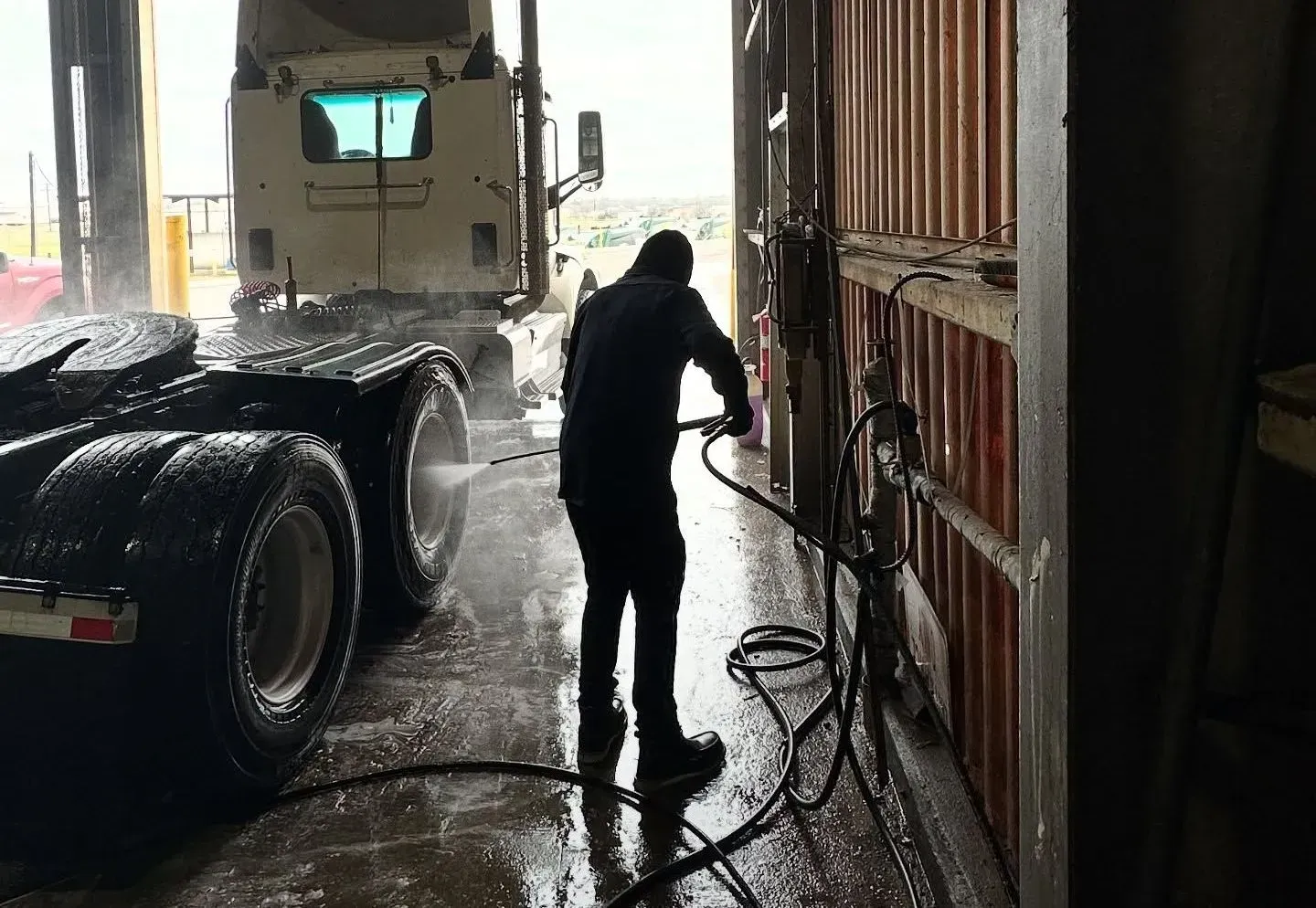 Worker pressure-washing a truck trailer inside a wet industrial bay