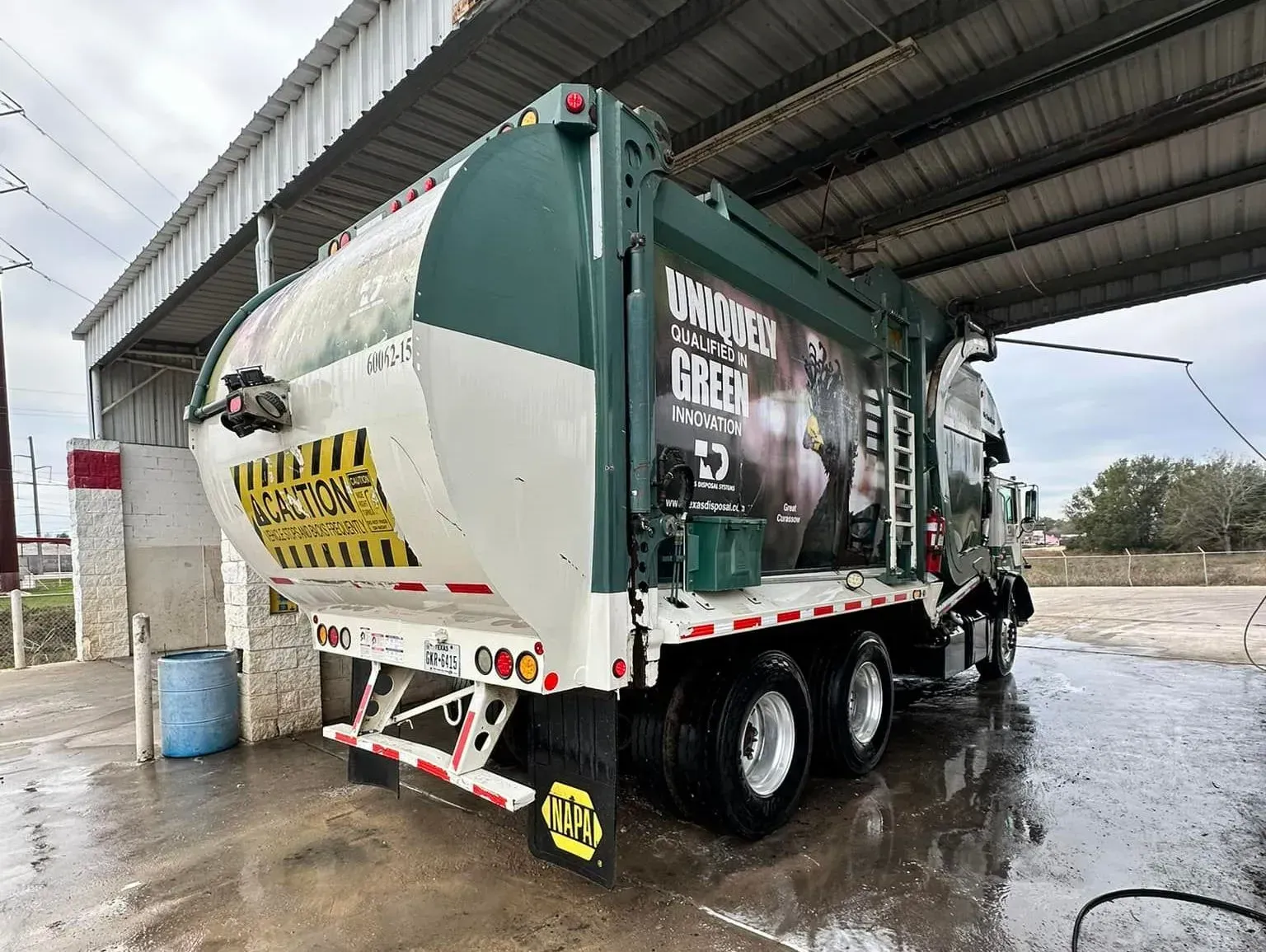 Green-and-white garbage truck at a wet fueling station under an overhang