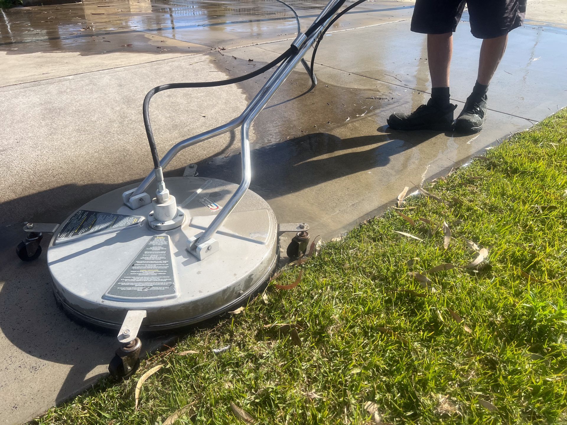 Person using a circular pressure washer on a wet sidewalk beside grass
