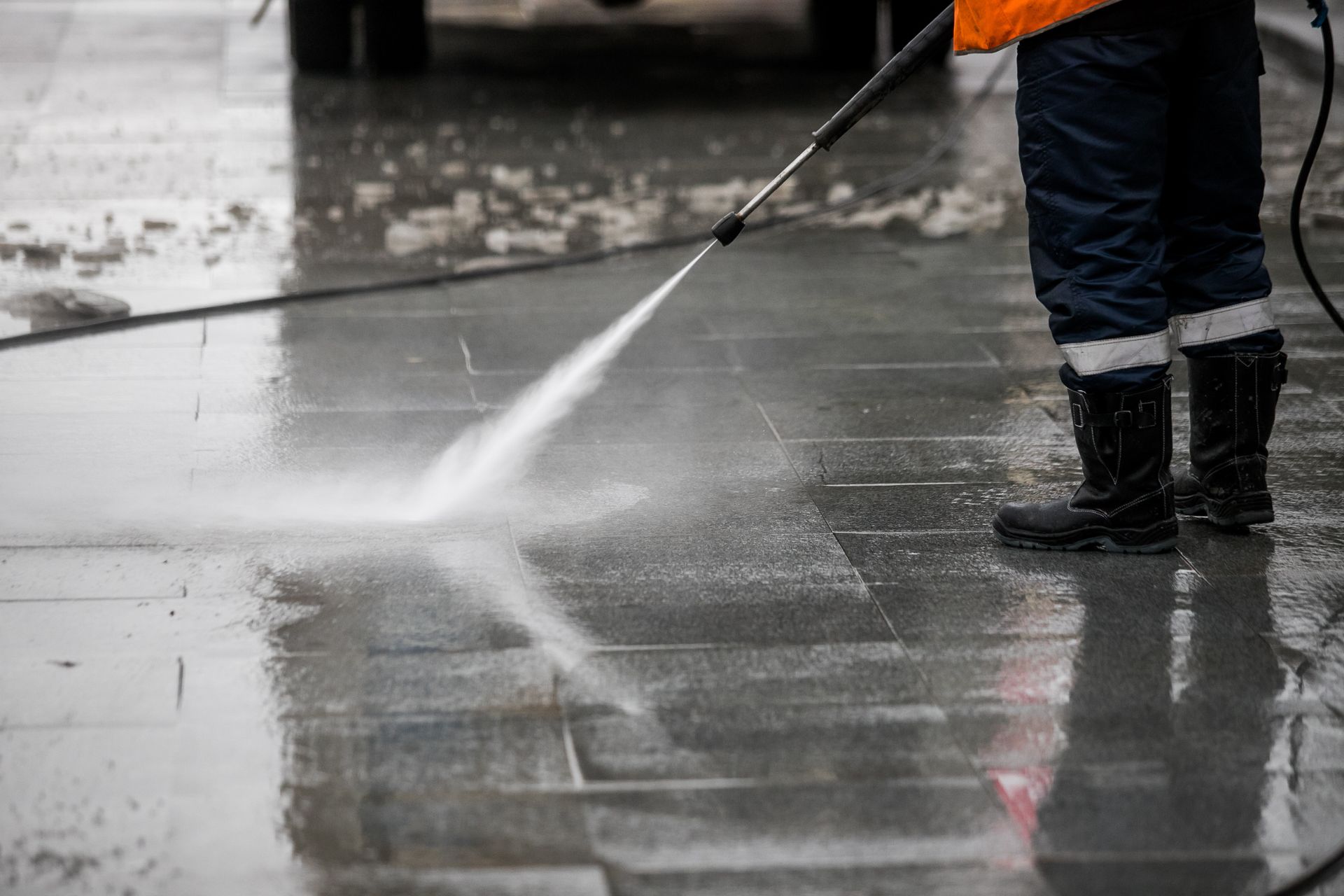 Person pressure-washing a wet sidewalk with a hose and spray wand, wearing black boots and orange jacket