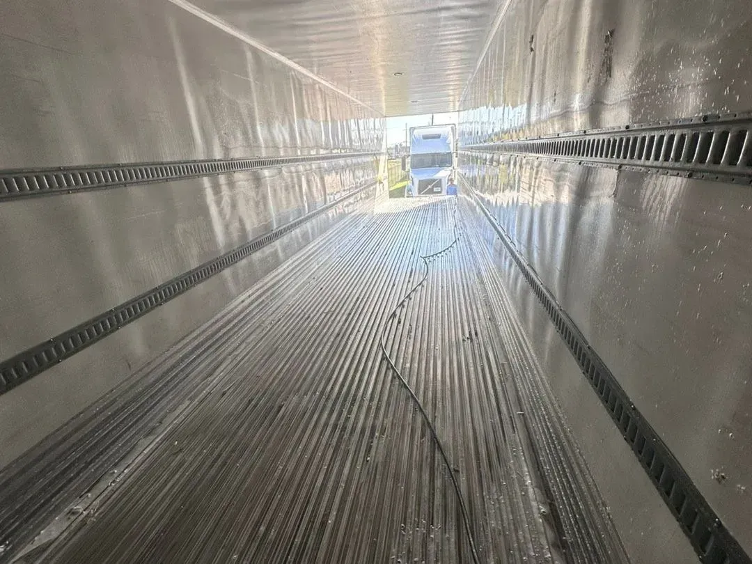 Empty metal cargo trailer with reflective walls and ribbed floor, viewed from the open rear toward the front.