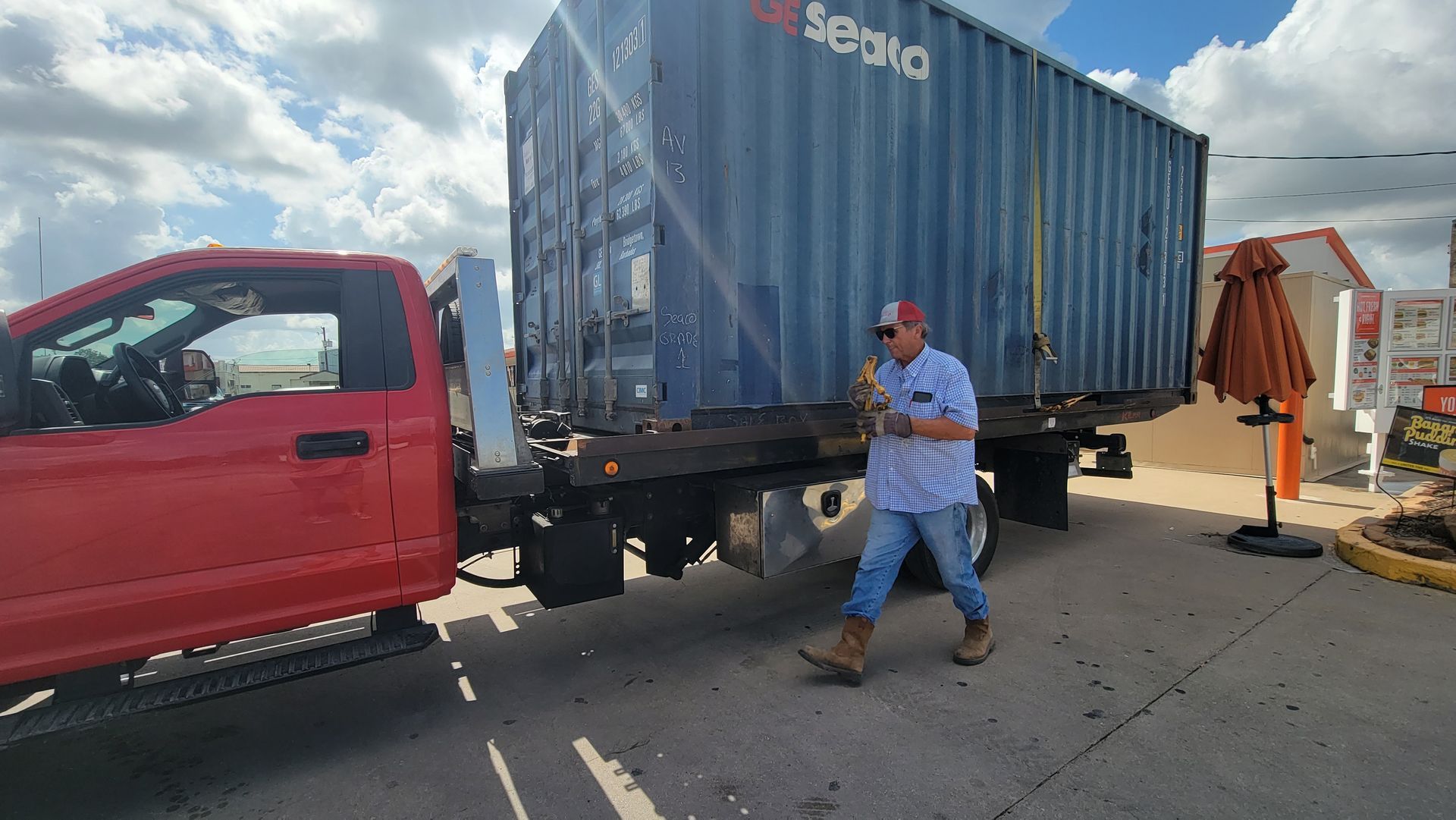 Worker standing beside a red truck carrying a blue shipping container under a cloudy sky