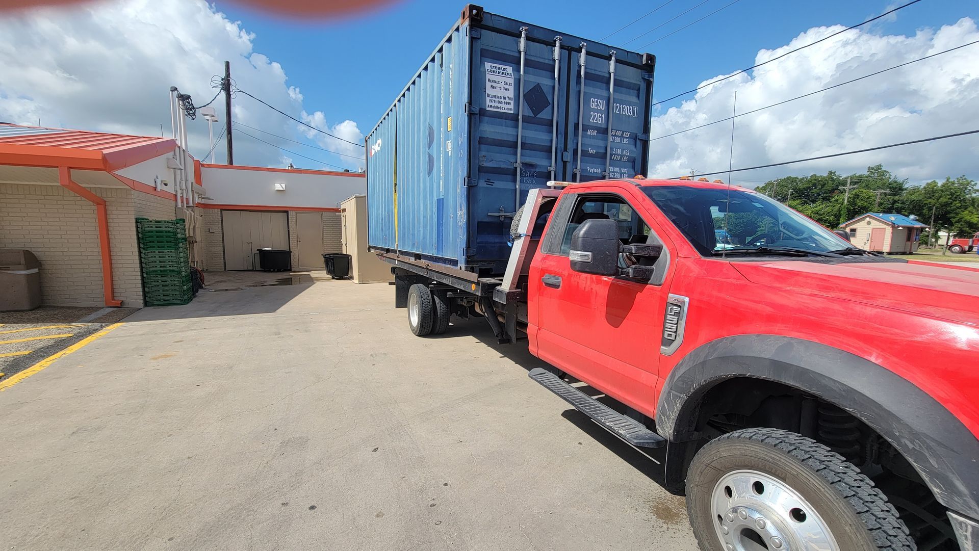 Red pickup truck hauling a blue shipping container in a sunny industrial yard